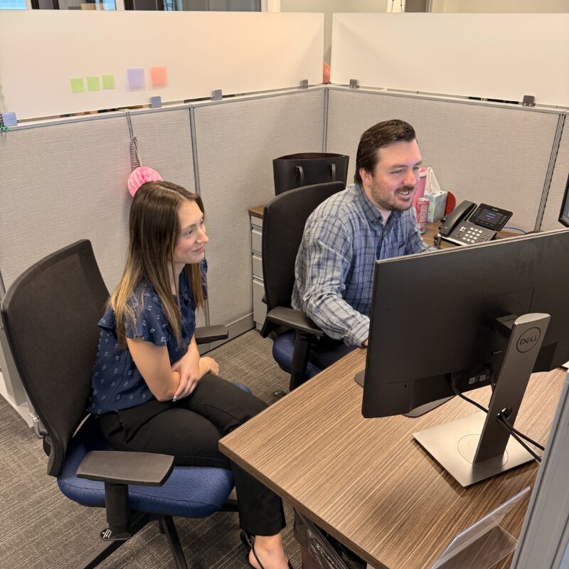 two coworkers site together looking at computer screens in a cubicle office